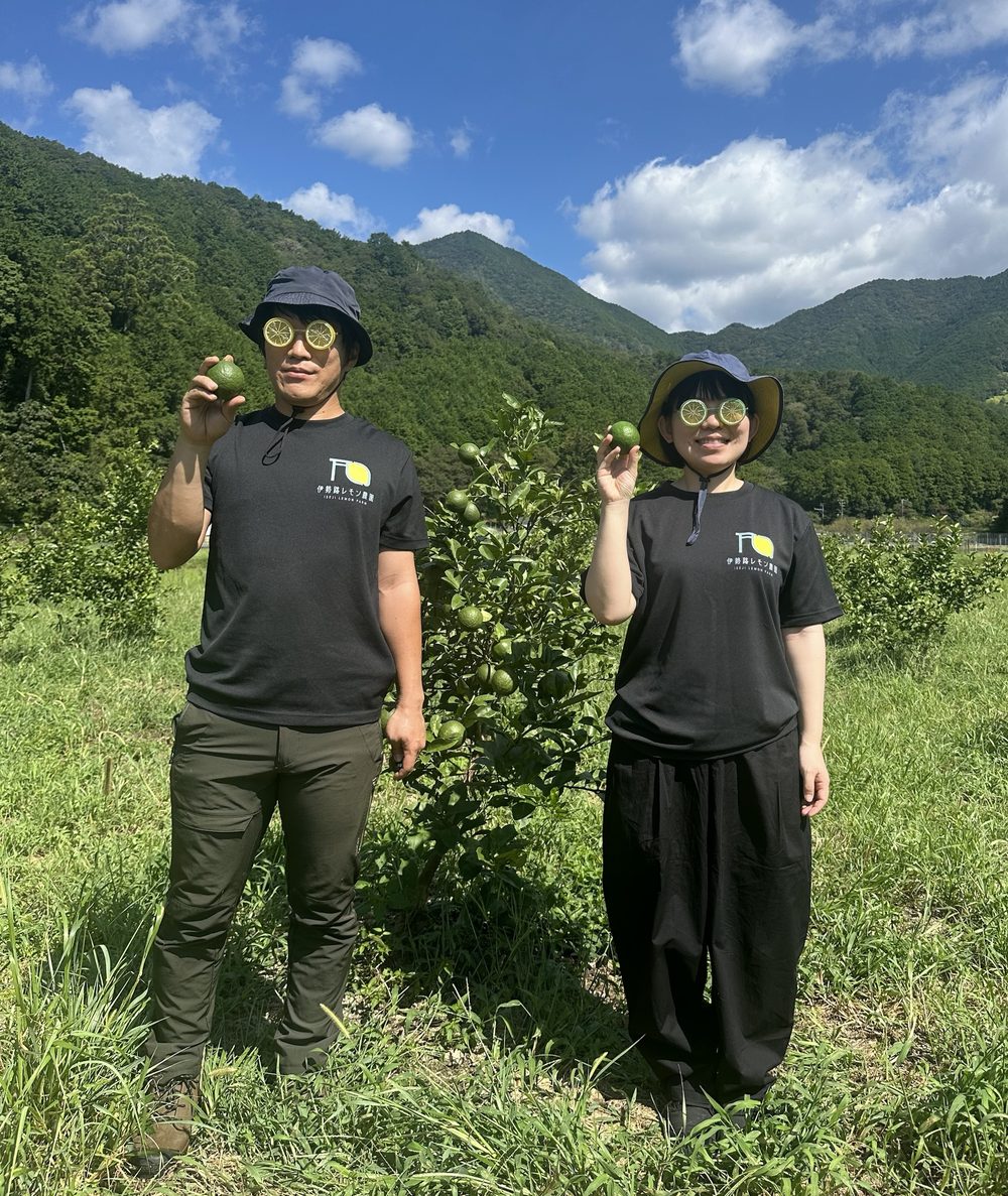 レモン眼鏡をかけ、青いレモンを手に畑に立つ園主夫婦。背景は紀北町の山々と青空